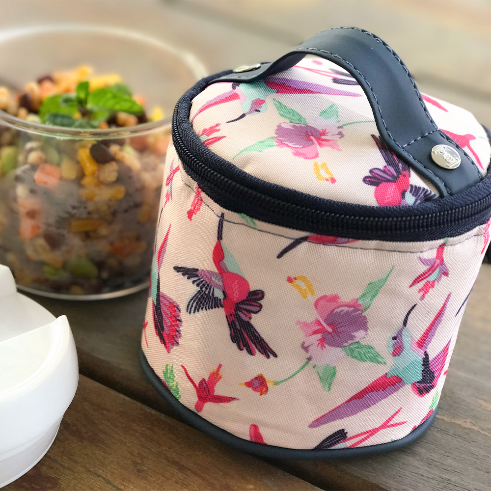 Bolsa térmica com estampa colorida de beija-flores sobre uma mesa de madeira, ao lado de um recipiente com salada de grãos.