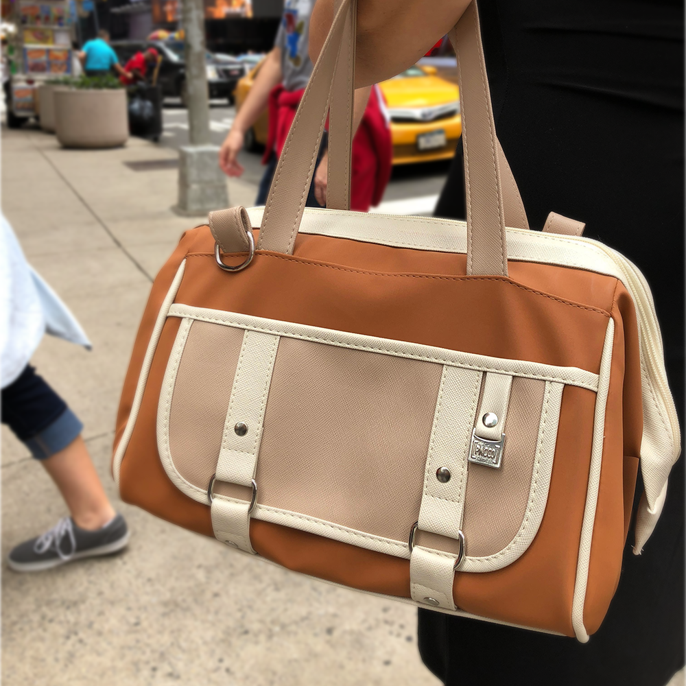 Pessoa carregando a bolsa térmica Vermont da PACCO na cor caramelo em uma calçada movimentada de uma grande cidade, com um táxi amarelo ao fundo.