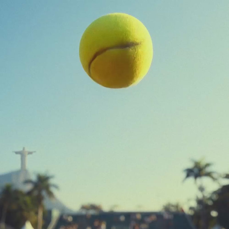 Vídeo da garrafa térmica Vyta Tennis PACCO com linhas brancas inspiradas na costura de uma bola de tênis. A garrafa é mostrada voando contra um céu azul limpo, sendo carregada por uma jogadora em uma quadra de saibro, guardada em uma bolsa de palha e posicionada ao lado de sua embalagem vermelha em um cenário do Rio Open.